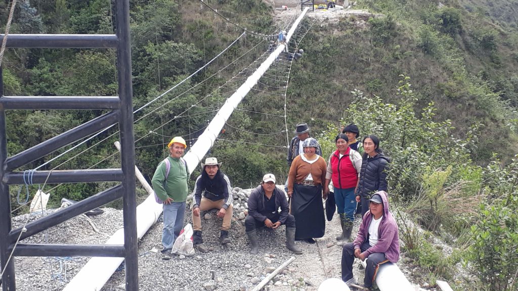 A group of community members stand near white pipe infrastructure that spans a ravine in Ecuador 